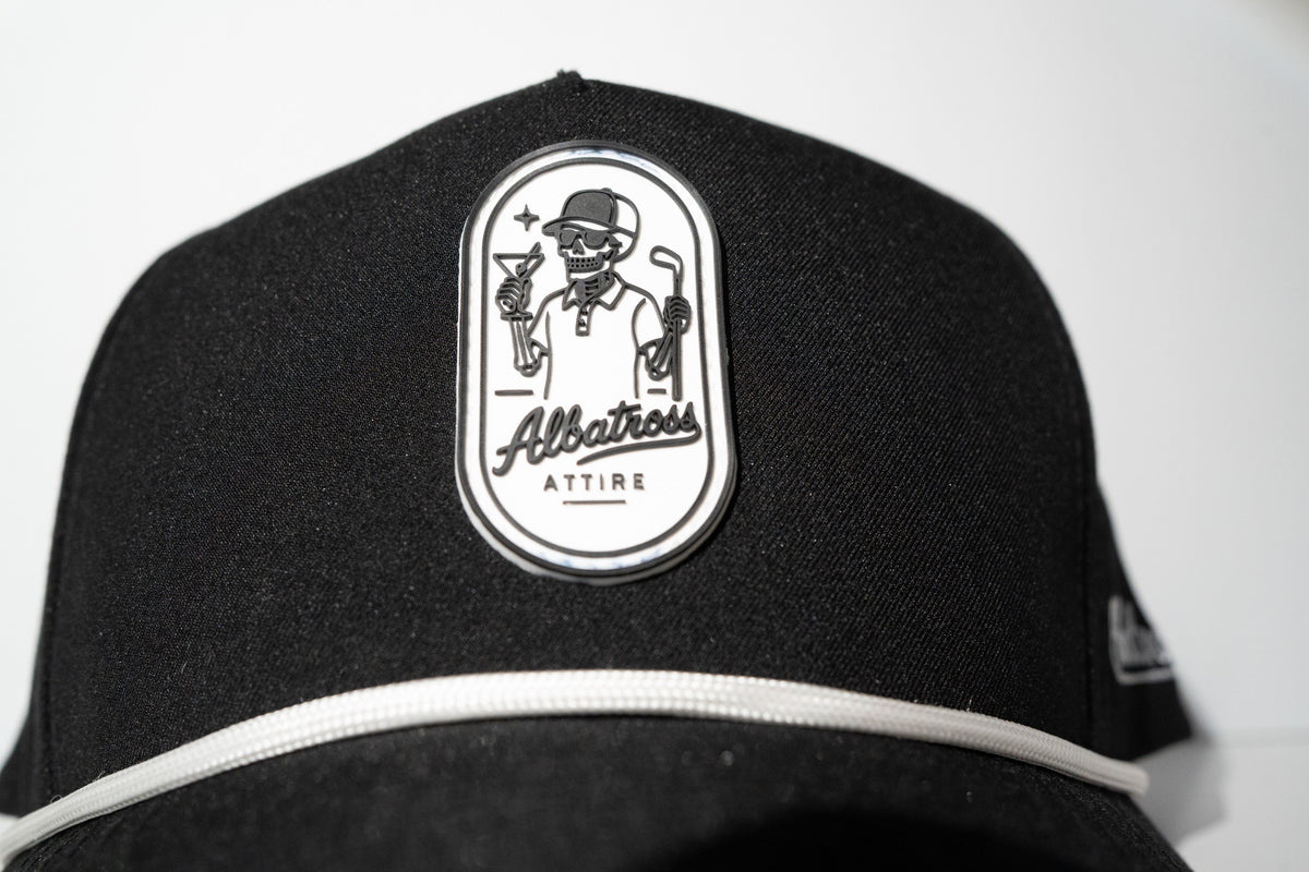 A close-up of a black baseball cap featuring a white embroidered oval patch on the front with a golf-themed illustration and the text 'Allatress ATTIRE'.