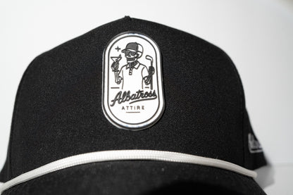 A close-up of a black baseball cap featuring a white embroidered oval patch on the front with a golf-themed illustration and the text 'Allatress ATTIRE'.