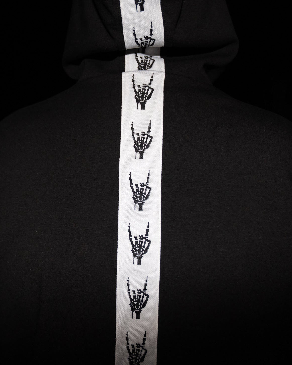 A black hoodie featuring a prominent white vertical stripe down the center, decorated with a repeating black graphic of skeletal hands making the rock on gesture.