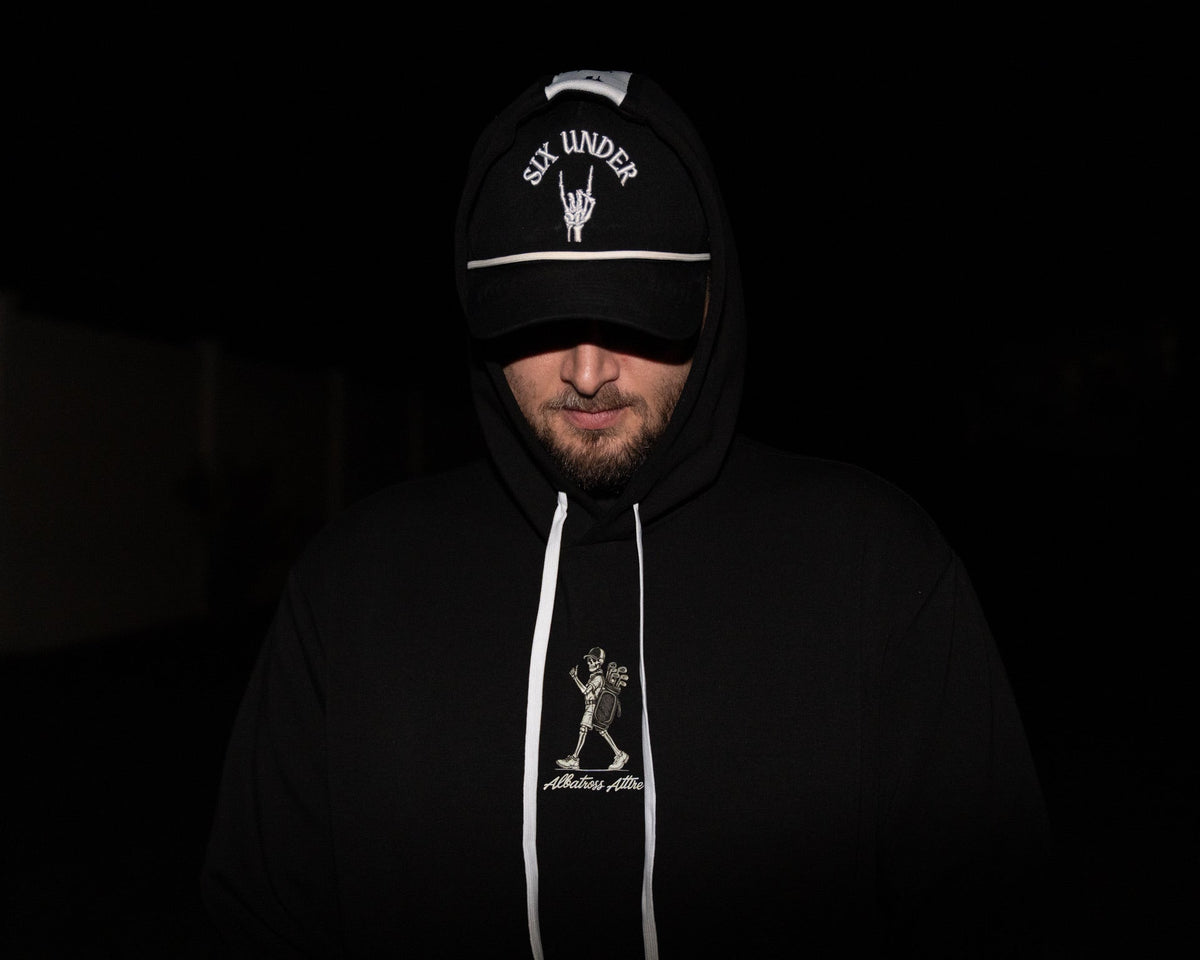 A man wearing a black hoodie and matching black cap with white graphics and text, photographed in very dark lighting against a nearly black background.
