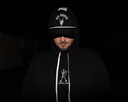 A man wearing a black hoodie and matching black cap with white graphics and text, photographed in very dark lighting against a nearly black background.