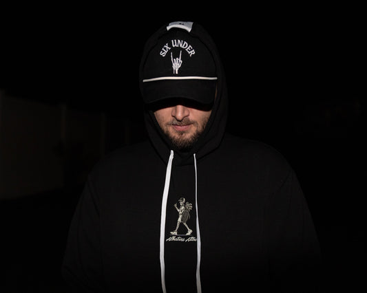 A man wearing a black hoodie and matching black cap with white graphics and text, photographed in very dark lighting against a nearly black background.